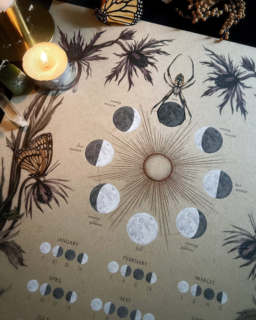 Decorative calendar with moon phases and botanical illustrations on a table with a candle.