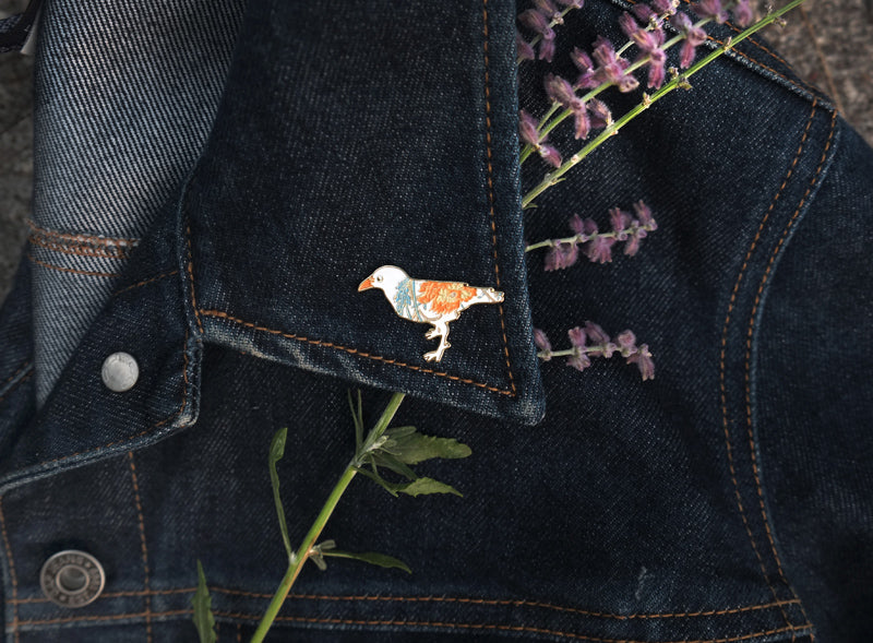 Small enamel pin of a white crow with orange and blue floral pattern.