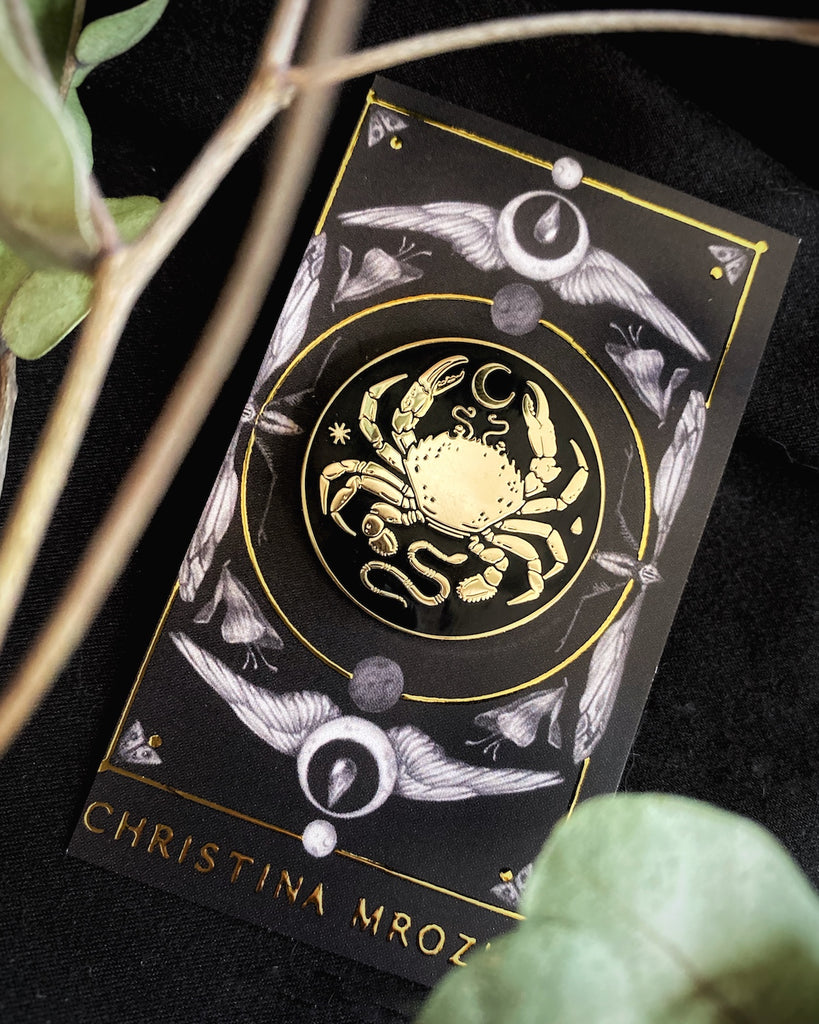 Small black enamel pin with gold-plated crab, moon, and snake design.