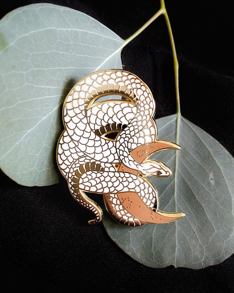 White enamel pin with gold plating of a snake wrapped around a clay-colored crescent moon.