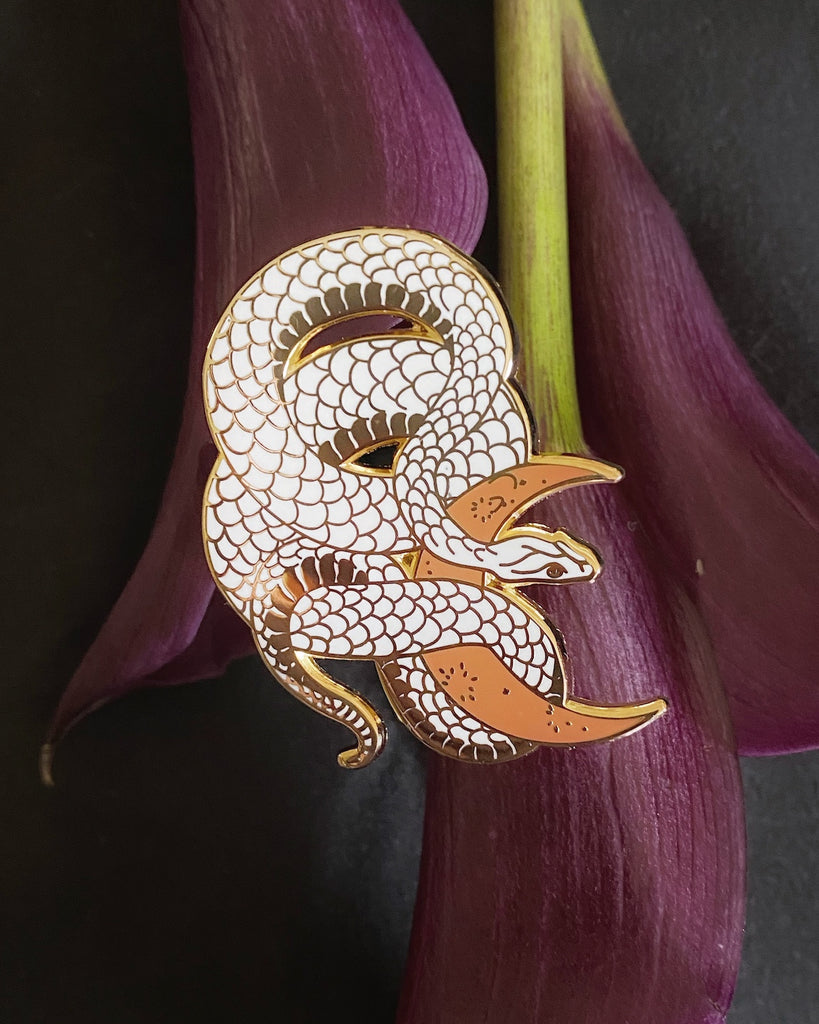 White enamel pin with gold plating of a snake wrapped around a clay-colored crescent moon.