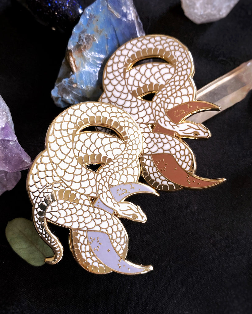 White enamel pin with gold plating of a snake wrapped around a crescent moon, shown in both lilac and clay.