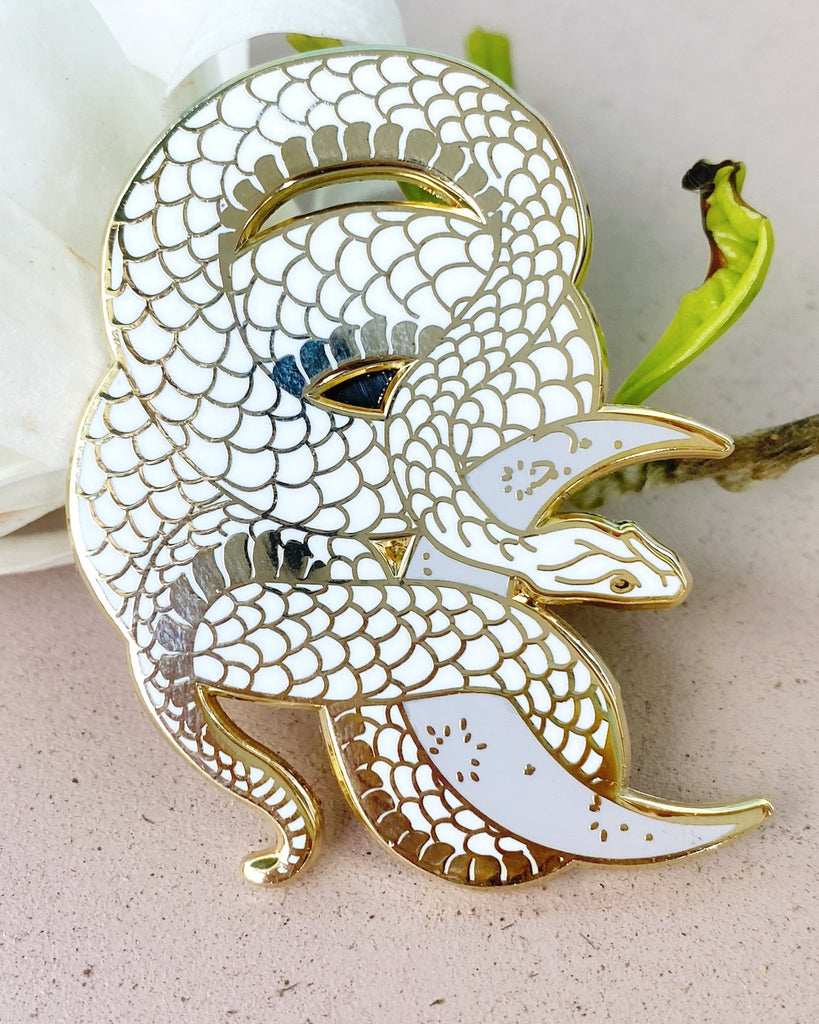 White enamel pin with gold plating of a snake wrapped around a lilac-colored crescent moon.