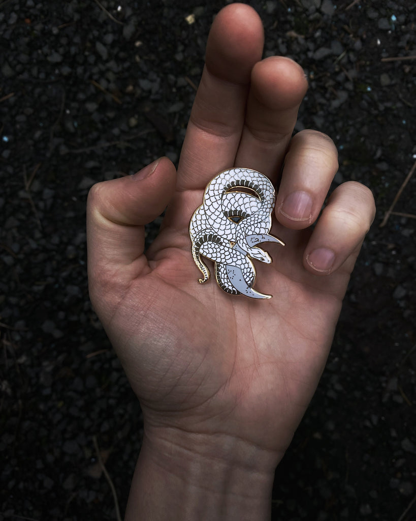 White enamel pin with gold plating of a snake wrapped around a lilac-colored crescent moon.