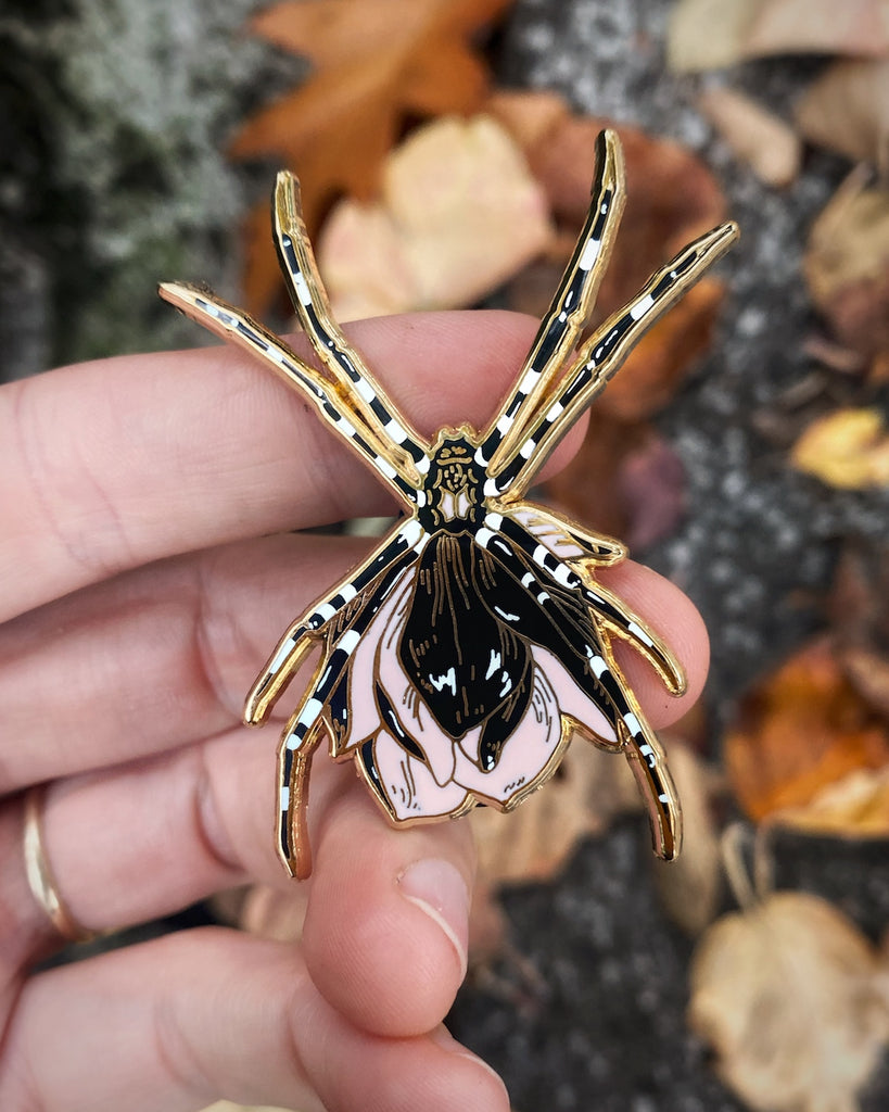 Gold plated enamel pin of a black orb weaver spider with a pink flower growing from its back.