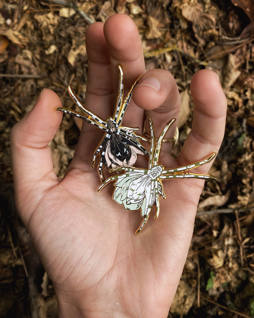 Gold plated enamel pin of a black orb weaver spider with a pink flower growing from its back, as well as an alternate color-way with white spider and green flower.