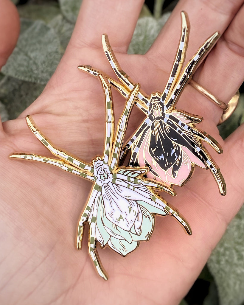 Gold plated enamel pin of a black orb weaver spider with a pink flower growing from its back, as well as an alternate color-way with white spider and green flower.. 