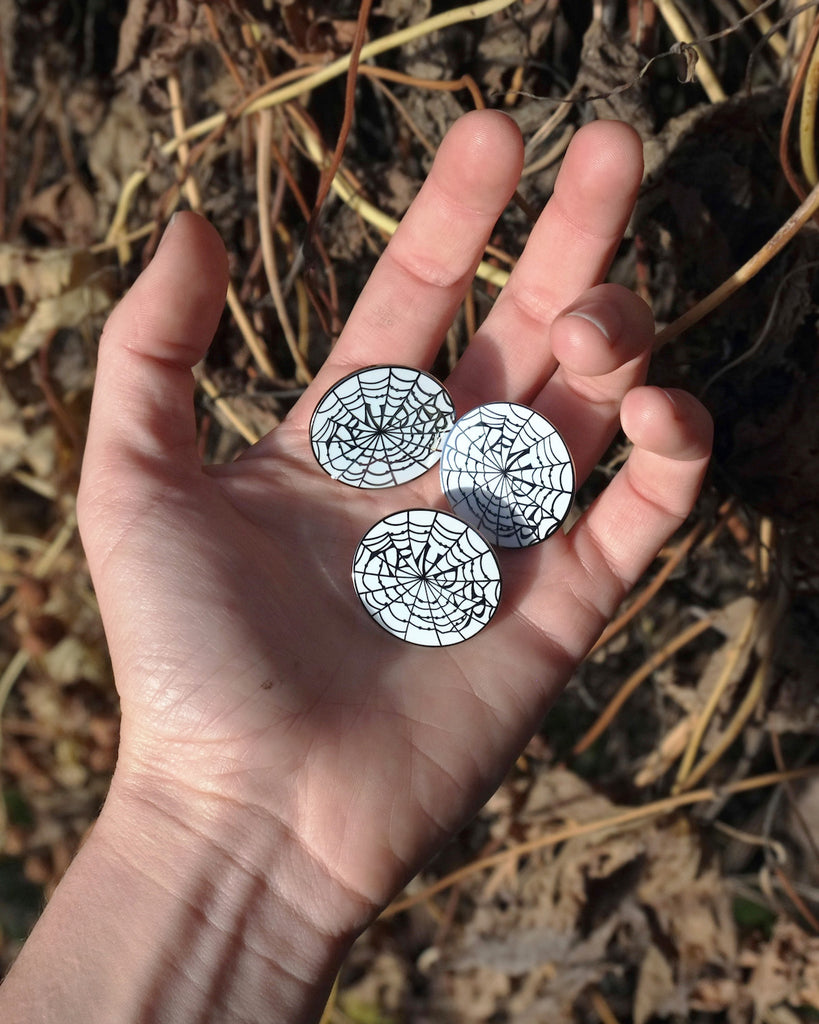 Small enamel pin with a white background and silver outline of a spider web that spells out “Tender”