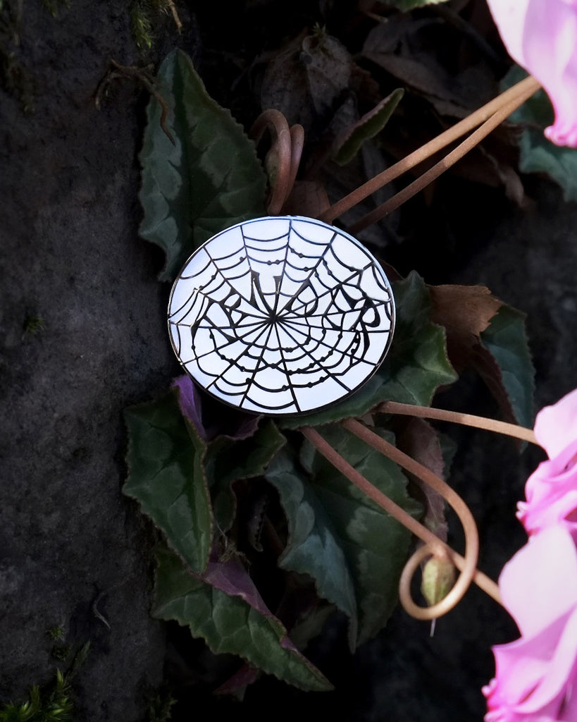 Small enamel pin with a white background and silver outline of a spider web that spells out “Tender”