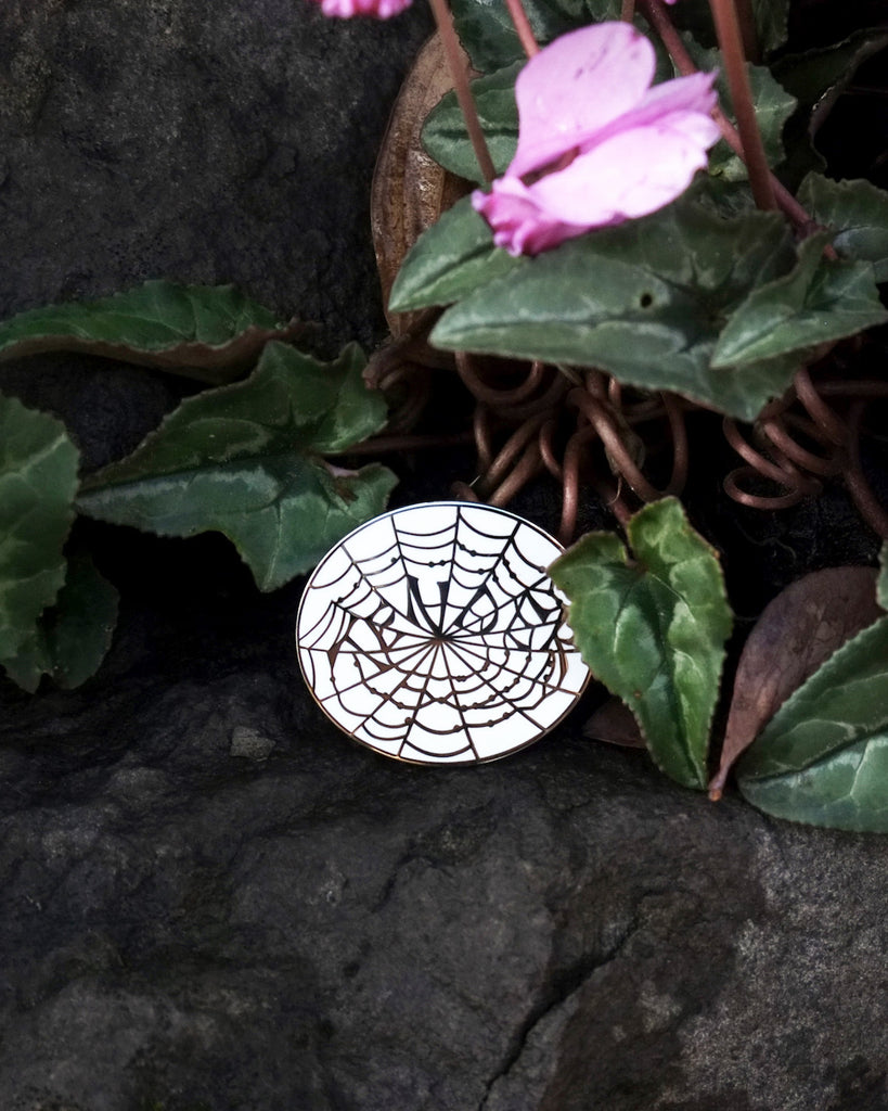 Small enamel pin with a white background and silver outline of a spider web that spells out “Tender”