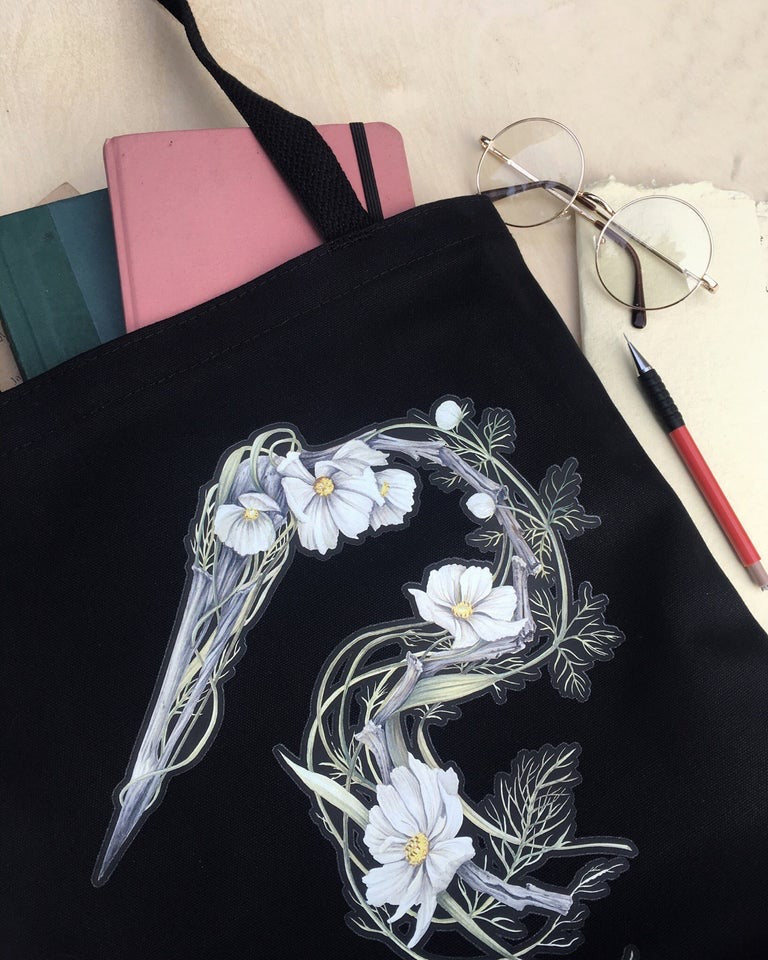 Black canvas tote bag featuring an illustration of a heron skeleton made of driftwood and white cosmo flowers.