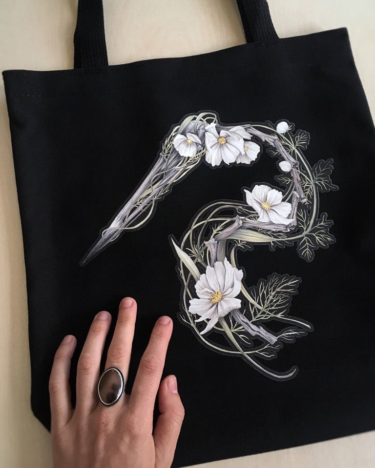 Black canvas tote bag featuring an illustration of a heron skeleton made of driftwood and white cosmo flowers.