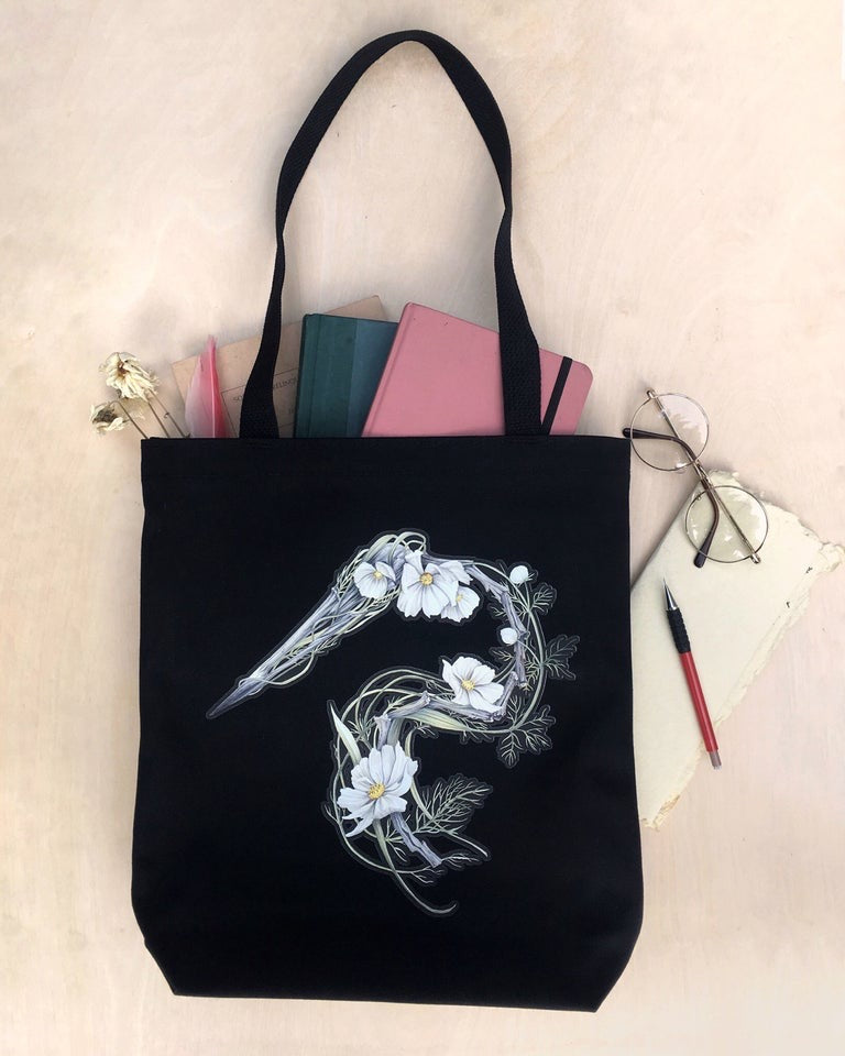 Black canvas tote bag featuring an illustration of a heron skeleton made of driftwood and white cosmo flowers.