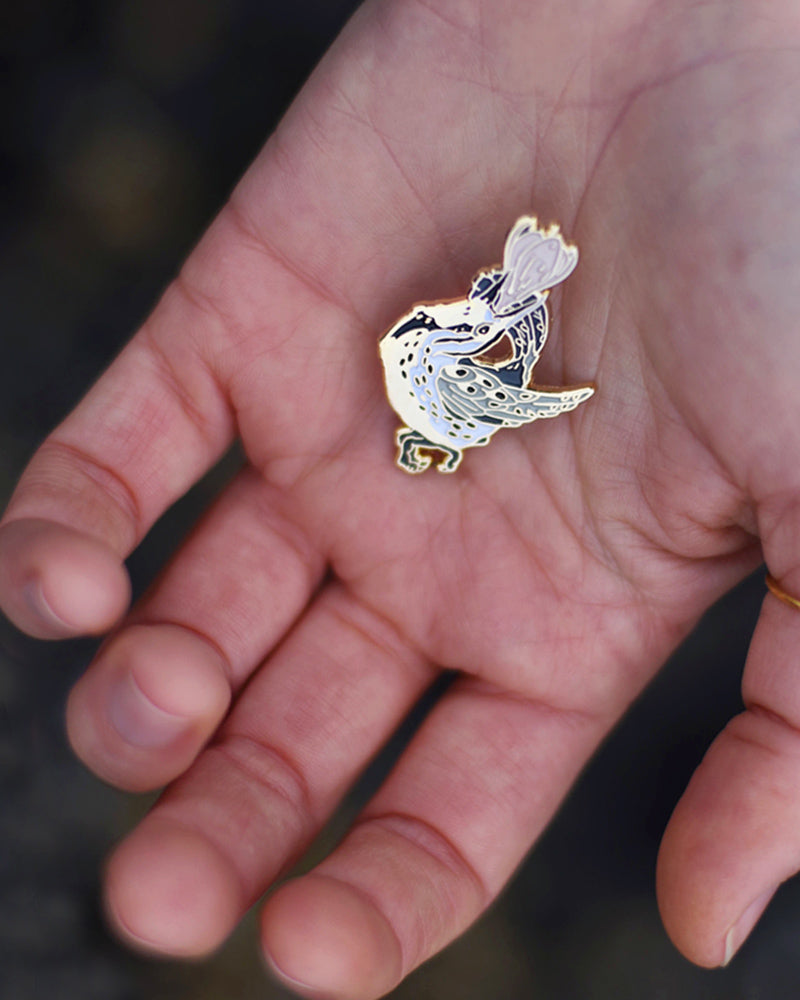 Small enamel pin of a black and white meadowlark with a pink flower growing from its open beak.