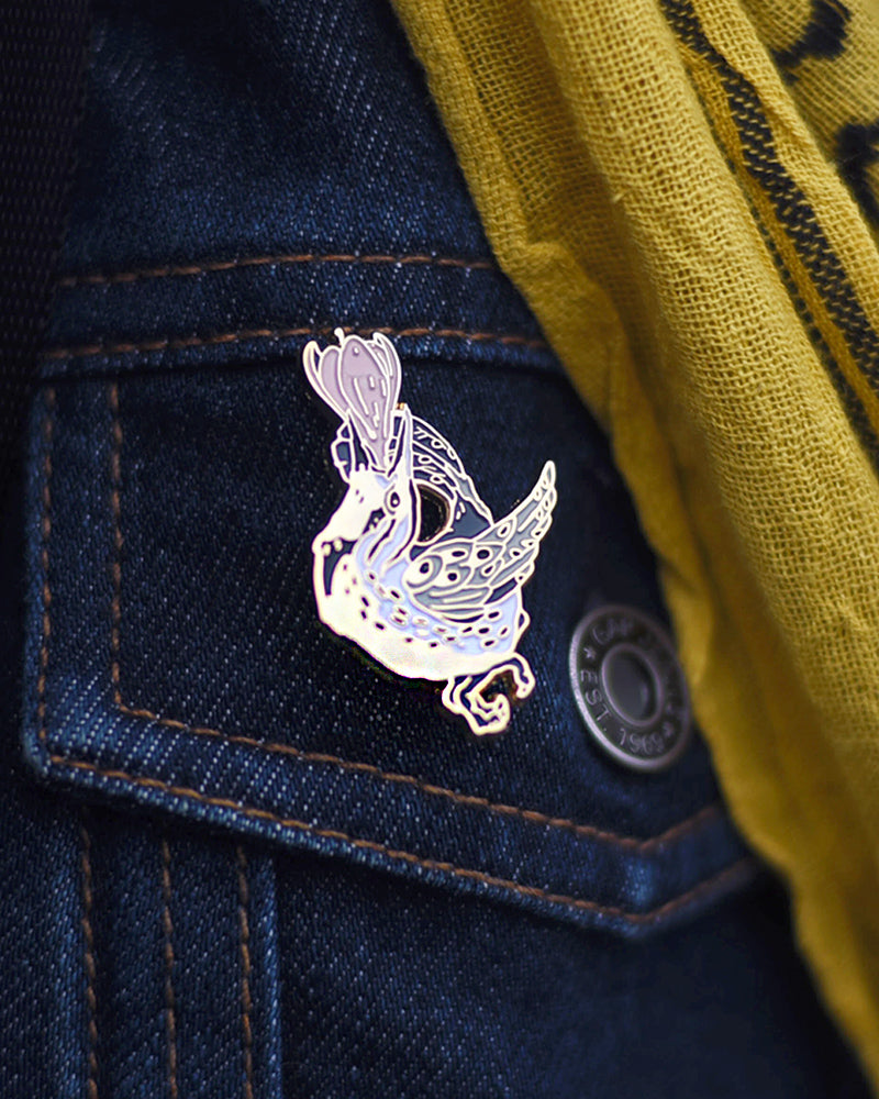 Small enamel pin of a black and white meadowlark with a pink flower growing from its open beak.