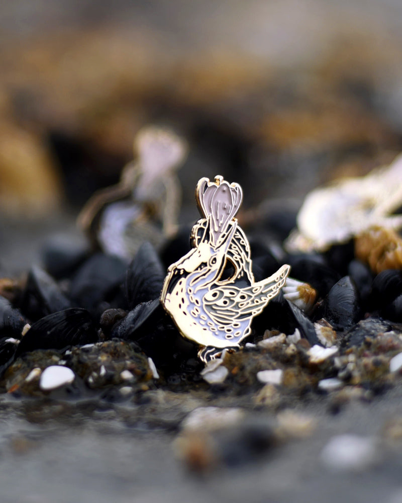 Small enamel pin of a black and white meadowlark with a pink flower growing from its open beak.