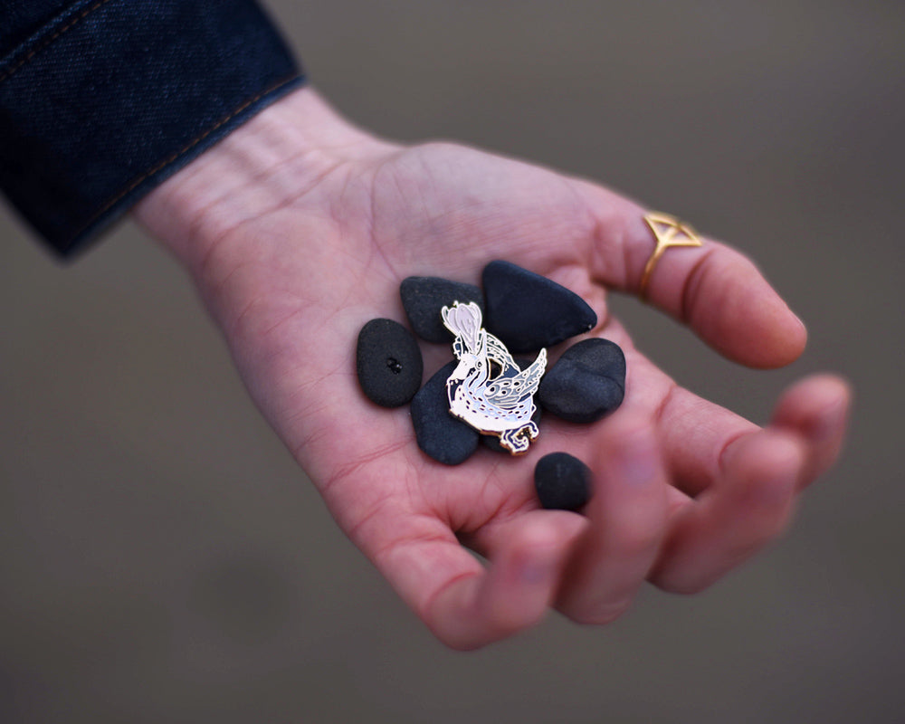 Small enamel pin of a black and white meadowlark with a pink flower growing from its open beak.